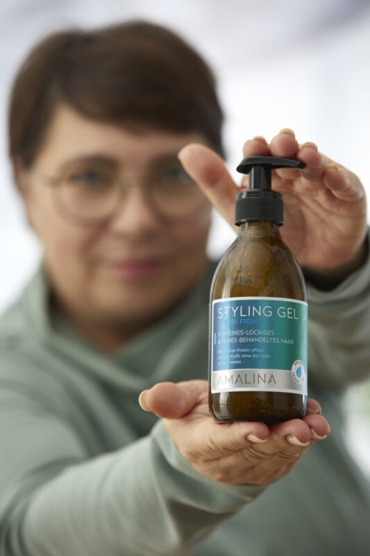 Eine Frau hält das Amalina Styling Gel in einer Glasflasche in die Kamera. Das Gel ist für feines, lockiges und behandeltes Haar geeignet und enthält pflanzliches Protein, das das Haar kräftigt.