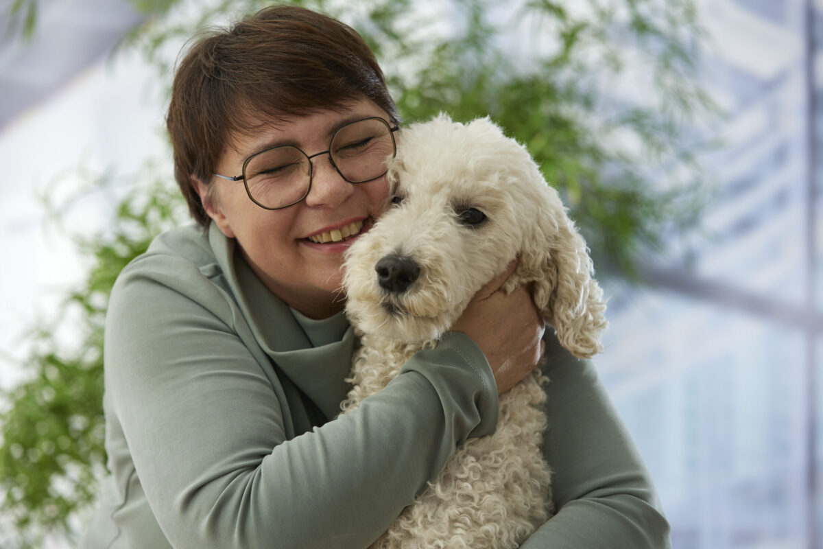 Eine Frau umarmt ihren Hund und lächelt. Sie trägt ein graues Oberteil, während der Hund mit lockigem, weißem Fell in ihren Armen sitzt. Der Hintergrund ist grün und vermittelt eine freundliche und entspannte Stimmung.